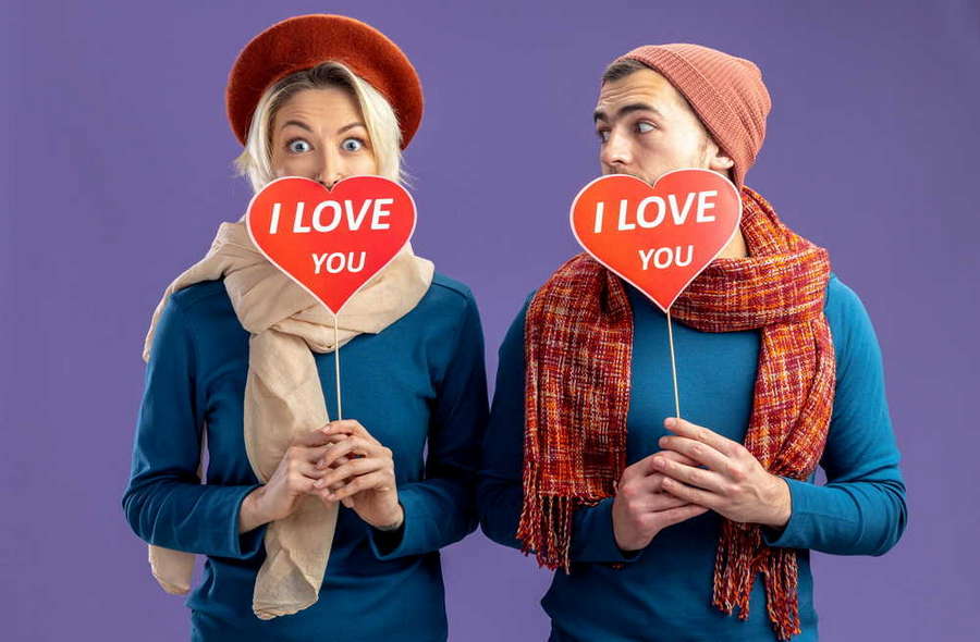 young couple wearing hat with scarf valentine s day covered face with red hearts stick with i love you text pleased guy looking girl isolated blue background